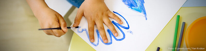 Webbanner Beitrag "Vorgaben". Motiv: Eine Kinderhand auf einem Blatt Papier, die andere Kinderhand malt mit blauer Wasserfarbe den Umriss der aufliegenden Hand nach.