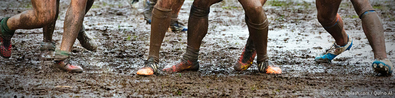 Banner "Andacht: Schlammschlacht": Eine Gruppe von Männern, die eine Partie Rugby spielen, man sieht nur die eingesauten Füße und Beine im Schlamm.